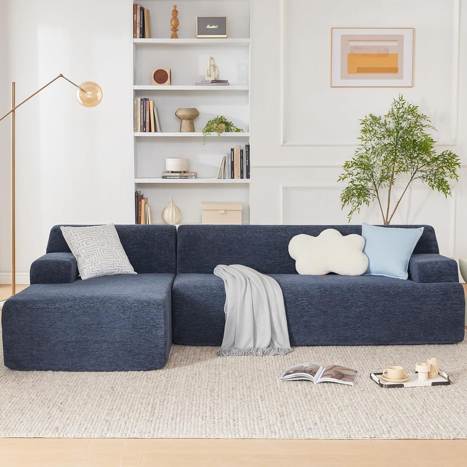 Front view: Navy blue chenille L-shaped sectional sofa slipcover in a modern minimalist living room, featuring decorative pillows, a grey throw blanket, a gold floor lamp, and a white built-in bookshelf background.